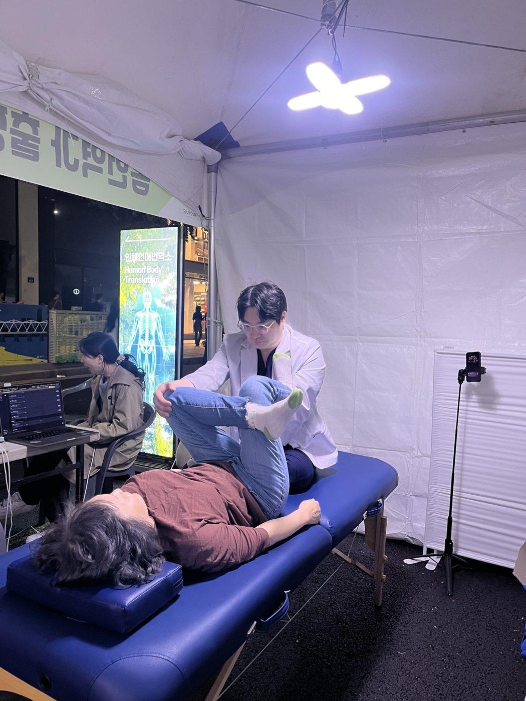 밤 축제 부스 안에서 물리치료사 김윤하가 누운 게스트의 다리를 들어 움직임을 살피는 장면